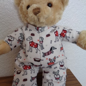 Puede incluir: Un oso de peluche beige con un pijama blanco estampado con perros rojos, grises y negros. El pijama presenta huesos y cuencos para comida. El oso tiene ojos oscuros y está sobre una superficie de madera.