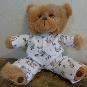 Pode incluir: Um urso de pelúcia marrom vestindo um pijama branco com estampa de coelhos, esquilos e flores. O urso tem um rosto sorridente e patas marrons. O pijama tem mangas compridas e calças.