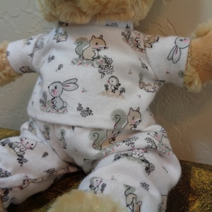 Pode incluir: Um urso de pelúcia vestindo um pijama branco com um padrão repetido de esquilos, coelhos e flores. O pijama tem mangas compridas e calças, com detalhes em rosa e cinza. O urso é marrom claro.