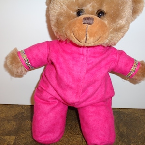 Pode incluir: Um urso de pelúcia marrom, vestindo um macacão rosa brilhante. O macacão tem faixas decorativas de strass coloridos nos punhos. O urso tem uma expressão sorridente e olhos escuros.