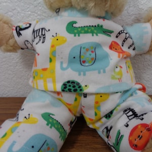 Pode incluir: Um urso de pelúcia vestindo um conjunto de pijama de duas peças. O pijama é branco com um padrão colorido de girafas, elefantes, crocodilos e outros animais. As mangas e punhos são azul claro.