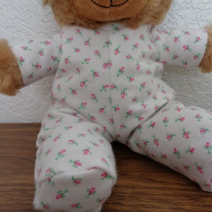 May include: A plush teddy bear wearing a white pajama set with a pink floral pattern. The bear has brown fur and is sitting on a wooden surface. The pajamas cover the bear's arms and legs.