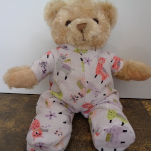 Puede incluir: Un oso de peluche con un mono blanco estampado con llamas coloridas, flores y formas geométricas. El oso tiene pelaje marrón claro y una expresión amigable. El mono presenta detalles en rosa, verde y morado.