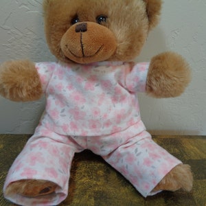 Pode incluir: Um urso de pelúcia marrom vestindo um pijama floral rosa e branco. O pijama apresenta um padrão de flores rosas e folhas cinzentas. O urso tem um rosto sorridente e está sentado com as pernas abertas.