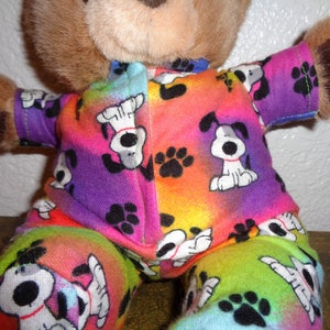 Pode incluir: Um urso de pelúcia marrom vestindo um pijama colorido. O pijama apresenta um padrão repetido de cães de desenho animado brancos e pretos e pegadas pretas em um fundo gradiente de arco-íris. O urso tem pelo marrom.