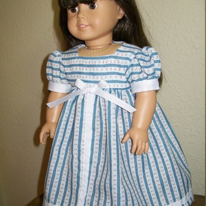 May include: A doll wearing a blue and white striped dress with a white ribbon bow at the waist. The dress has short sleeves and a white ribbon trim at the hem. The doll has brown hair and brown eyes.