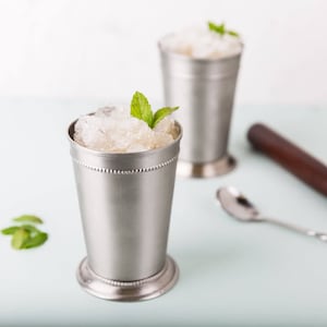 May include: Two silver metal cups filled with crushed ice and a mint garnish. The cups are on a light blue surface.