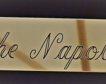 Puede incluir: Un letrero de metal de color dorado con esquinas redondeadas y un borde decorativo. El letrero dice "~ The Napoleon ~" en una fuente cursiva.