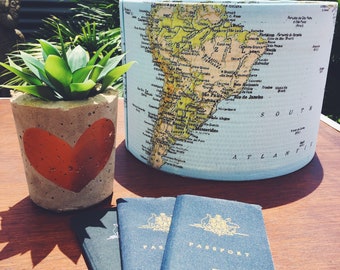May include: A light blue lampshade with a map of South America printed on it. The lampshade is sitting on a wooden table next to a concrete planter with a red heart design and two blue passports with gold lettering that says "PASSPORT".