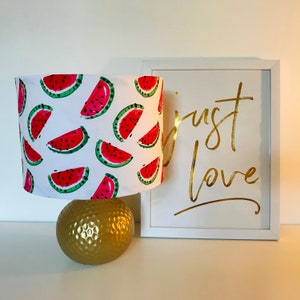 May include: A white lampshade with a watermelon print. The lampshade is on a gold base. A white frame with the text "just love" in gold lettering is in the background.