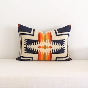 May include: Rectangular decorative pillow with a geometric pattern in cream, navy blue, orange, and brown. The pillow is placed on a white bench, showcasing its design and colors. The pillow's design features a central diamond shape.