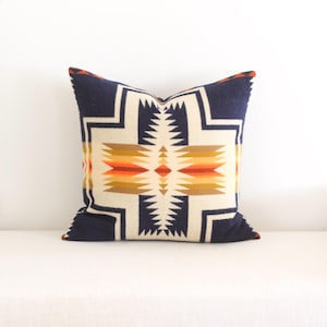 May include: Square decorative pillow with a geometric pattern in navy blue, cream, orange, and brown. The pillow features a central design with a symmetrical arrangement of shapes and colors, suitable for home decor.