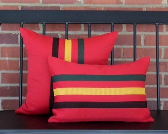 May include: Two red throw pillows with striped accents in black, yellow, and green sit on a black bench. One pillow is rectangular, and the other is square. The pillows are a decorative accent for a home.