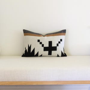 May include: Rectangular cushion with a geometric pattern. The cushion has a cream-coloured background with a black cross and black triangular shapes at the bottom. It has a grey and brown stripe at the top. The cushion is on a white bench.