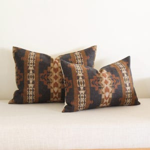 May include: Two rectangular throw cushions featuring a geometric design in brown, tan, and grey. The cushions are placed on a white surface, suitable for home decor.
