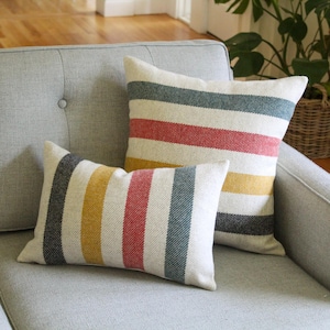 May include: Two decorative throw pillows with a striped pattern in red, yellow, blue, and black on a cream background. The pillows are placed on a gray sofa, creating a cozy and inviting atmosphere. The pillows add a pop of color and texture to the living space.