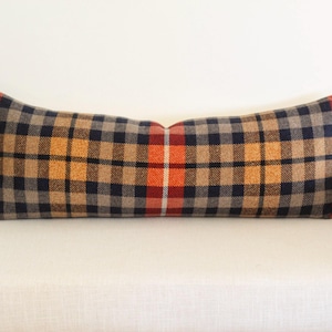 May include: A long rectangular pillow with a plaid pattern in shades of navy, tan, orange, and red. The pillow is placed on a white surface, suggesting a decorative accent for a sofa or bed. The plaid design adds a touch of rustic charm.