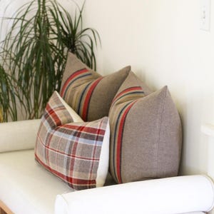 May include: Three decorative throw pillows on a white bench. Two pillows are a solid brown with red and blue stripes. The third pillow has a plaid pattern in red, brown, and blue. A plant is in the background.