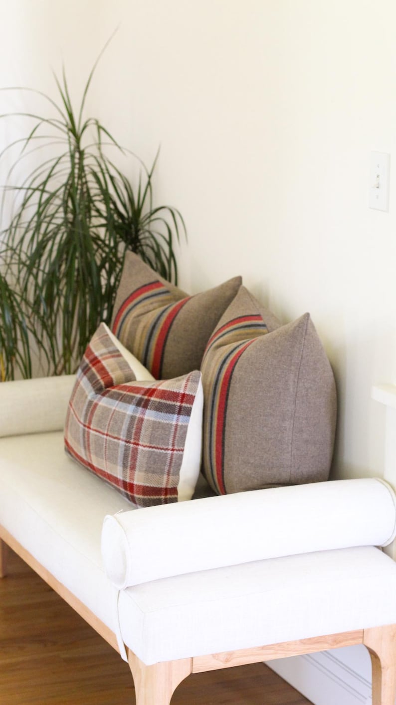 May include: Three decorative throw pillows on a white bench. Two pillows are a solid brown with red and blue stripes. The third pillow has a plaid pattern in red, brown, and blue. A plant is in the background.