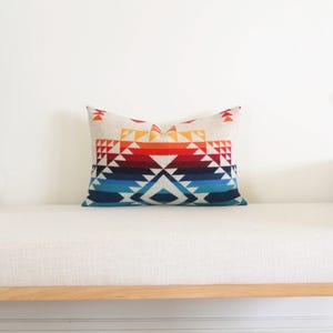 May include: A rectangular cushion featuring a geometric design in red, orange, yellow, blue, and white. The cushion is placed on a white bench with a wooden frame.