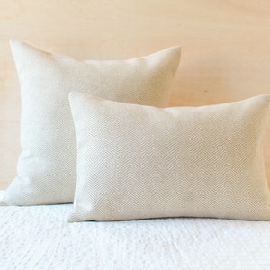 May include: Two beige throw pillows with a subtle herringbone pattern. The pillows are arranged on a white bedspread.