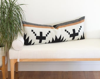 May include: A long, rectangular decorative pillow with a geometric pattern. The pillow features a white background with black, brown, and gray accents. The design includes black crosses and jagged lines. The pillow is resting on a white bench.