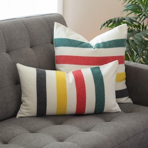 May include: Two cushions with a striped pattern in red, green, yellow, and grey on a white background. The cushions are placed on a grey tufted sofa. The cushions add a pop of colour to the neutral-toned living room.