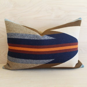 May include: Rectangular cushion with a geometric pattern. The cushion features a navy blue, rust, and orange stripe design, with tan and brown accents. The cushion is placed on a light wood surface.