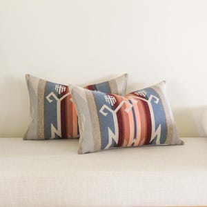 May include: Two rectangular throw pillows with a Southwestern design. The pillows display a geometric pattern in blue, beige, red, and orange. They are placed on a white bench.