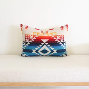 May include: A rectangular cushion with a geometric pattern in red, orange, blue, and white. The cushion is placed on a white bench. The design features triangles and diamond shapes. The cushion measures approximately 40cm x 60cm.