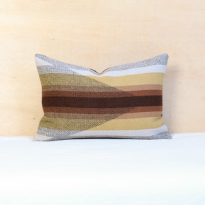 May include: Rectangular cushion with a geometric pattern. It features horizontal stripes in shades of brown, tan, and cream, with a diagonal section of darker brown. The cushion is placed on a white surface.