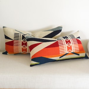 May include: Two rectangular cushions with a geometric pattern in cream, orange, red, navy, and white. The cushions are placed on a white sofa. The design features stripes and shapes.