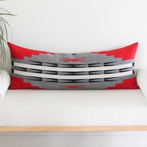 May include: A rectangular decorative pillow with a red border and a geometric pattern in gray, black, and white. The pillow is placed on a white bench with cylindrical bolsters on each side. The pillow's design features a series of stepped lines.
