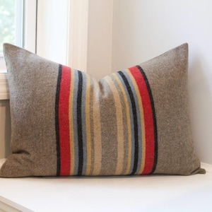 May include: A rectangular pillow with a brown wool exterior. The pillow features a central vertical stripe design with red, yellow, gray, and black stripes. The pillow is resting on a white surface near a window.