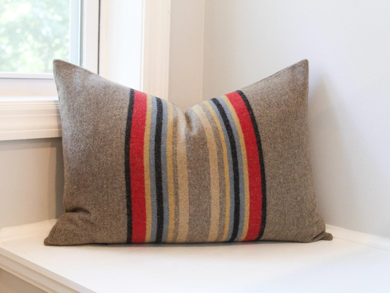 May include: A rectangular pillow with a brown wool exterior. The pillow features a central vertical stripe design with red, yellow, gray, and black stripes. The pillow is resting on a white surface near a window.