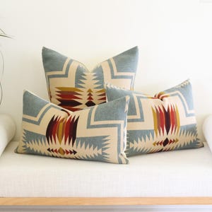 May include: Three decorative throw pillows with a geometric pattern in shades of blue, beige, red, and yellow. The pillows are arranged on a white bench with a wooden frame, creating a cozy and stylish look for home decor.