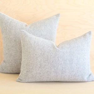 May include: Two decorative throw pillows with a grey and white herringbone pattern. The pillows are arranged on a light wood surface.