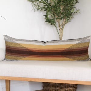May include: A long, rectangular decorative pillow with a geometric pattern in shades of gray, yellow, and brown. The pillow is placed on a white bench with a wooden frame. The pillow's design features angled stripes.