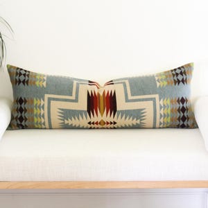 May include: A long rectangular lumbar pillow featuring a geometric pattern in shades of light blue, cream, dark brown, red, and yellow. The design consists of repeating triangles, diamonds, and stepped lines centered on the pillow face.