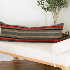 May include: A long, rectangular pillow with a striped pattern in shades of green, red, yellow, and black. The pillow is resting on a white bench with a wooden frame.