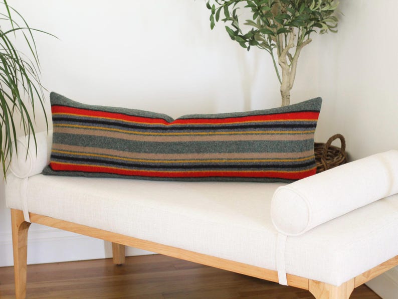 May include: A long, rectangular pillow with a striped pattern in shades of green, red, yellow, and black. The pillow is resting on a white bench with a wooden frame.