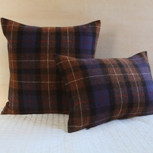 May include: Two plaid throw pillows, one larger than the other, with a brown, tan, and blue plaid pattern. The pillows are on a white bedspread.
