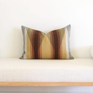 May include: Rectangular cushion with a geometric pattern in shades of brown, tan, and grey. The cushion has a textured appearance and is placed on a white bench with wooden legs. The design features vertical stripes.