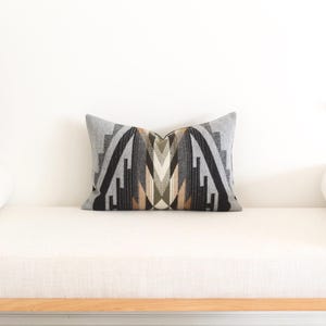 May include: Rectangular cushion with a geometric pattern in shades of grey, black, and beige. The cushion is placed on a white bench with a wooden frame, against a white wall. The cushion's design features a symmetrical arrangement of shapes.