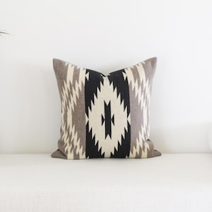May include: Square decorative pillow with a geometric pattern in black, gray, and cream colors. The pillow is placed on a white surface, showcasing its design and texture. The pillow's design features a central diamond shape.