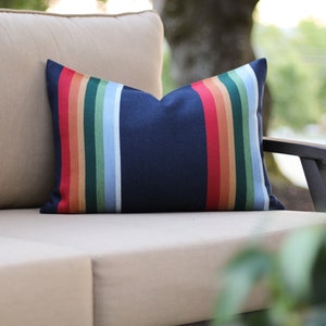 May include: Navy blue rectangular pillow with vertical stripes in red, orange, tan, green, light blue, and white. The pillow is placed on a beige outdoor sofa. The pillow's design adds a pop of color to the neutral setting.