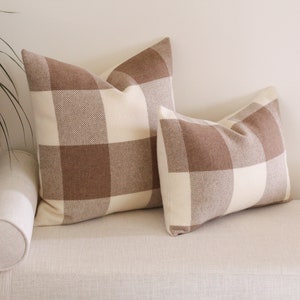 May include: Two brown and white plaid throw cushions on a cream coloured sofa. The larger cushion is in the background and the smaller cushion is in the foreground.