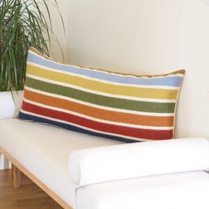 May include: A rectangular pillow with horizontal stripes in various colors, including blue, yellow, green, red, and orange. The pillow is placed on a white bench with rounded bolster pillows on each end.