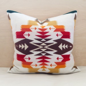 May include: A white pillow with a geometric pattern in shades of red, orange, brown, and green. The pattern is symmetrical and features arrows and triangles.