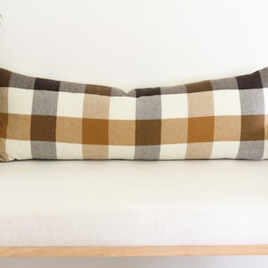 May include: A long rectangular lumbar cushion featuring a large-scale plaid pattern. The fabric displays a repeating grid design in alternating blocks of cream, warm tan, dark brown, and soft grey, creating a balanced and structured textile appearance.